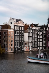 Several traditional Dutch canal houses with steep gabled roofs line up along a calm waterway. A boat labeled 'Canal Tours Amsterdam' is floating on the water, and a few people are seen near the edge of the canal. The sky is lightly overcast, adding a serene atmosphere.