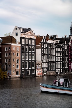 Several traditional Dutch canal houses with steep gabled roofs line up along a calm waterway. A boat labeled 'Canal Tours Amsterdam' is floating on the water, and a few people are seen near the edge of the canal. The sky is lightly overcast, adding a serene atmosphere.