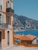 A coastal Mediterranean scene features a peach-colored building with ornate iron balconies. Two people are standing on a cobblestone path beside the building, looking toward a stunning sea view. The vibrant blue water is dotted with white yachts, and lush green mountains rise in the background.