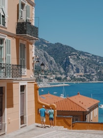 A coastal Mediterranean scene features a peach-colored building with ornate iron balconies. Two people are standing on a cobblestone path beside the building, looking toward a stunning sea view. The vibrant blue water is dotted with white yachts, and lush green mountains rise in the background.