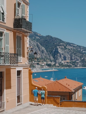 A coastal Mediterranean scene features a peach-colored building with ornate iron balconies. Two people are standing on a cobblestone path beside the building, looking toward a stunning sea view. The vibrant blue water is dotted with white yachts, and lush green mountains rise in the background.