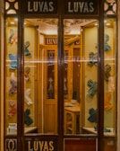 A quaint shop window display featuring an array of colorful leather gloves neatly arranged on a wooden background. The interior is warmly lit, showcasing the elegance and variety of the gloves. The vintage aesthetic of the store is enhanced by the decorative glass panels and wooden frames.