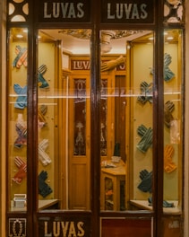 A quaint shop window display featuring an array of colorful leather gloves neatly arranged on a wooden background. The interior is warmly lit, showcasing the elegance and variety of the gloves. The vintage aesthetic of the store is enhanced by the decorative glass panels and wooden frames.