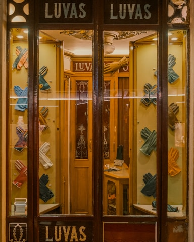 A quaint shop window display featuring an array of colorful leather gloves neatly arranged on a wooden background. The interior is warmly lit, showcasing the elegance and variety of the gloves. The vintage aesthetic of the store is enhanced by the decorative glass panels and wooden frames.