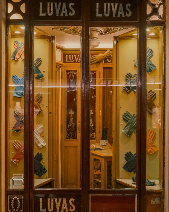 A quaint shop window display featuring an array of colorful leather gloves neatly arranged on a wooden background. The interior is warmly lit, showcasing the elegance and variety of the gloves. The vintage aesthetic of the store is enhanced by the decorative glass panels and wooden frames.