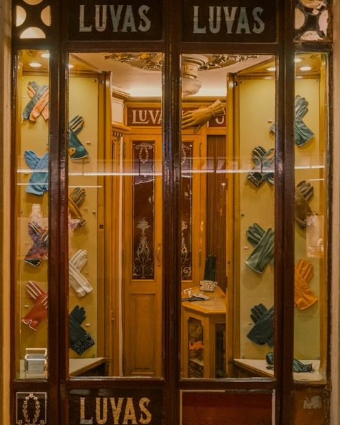 A quaint shop window display featuring an array of colorful leather gloves neatly arranged on a wooden background. The interior is warmly lit, showcasing the elegance and variety of the gloves. The vintage aesthetic of the store is enhanced by the decorative glass panels and wooden frames.