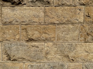 A close-up view of a stone wall with large, rectangular blocks. The stones are unevenly textured, showcasing natural variations in color and form. Each block has a slightly different hue, ranging from light tan to deeper brown, with visible mortar lines between the stones.