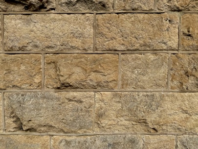 A close-up view of a stone wall with large, rectangular blocks. The stones are unevenly textured, showcasing natural variations in color and form. Each block has a slightly different hue, ranging from light tan to deeper brown, with visible mortar lines between the stones.