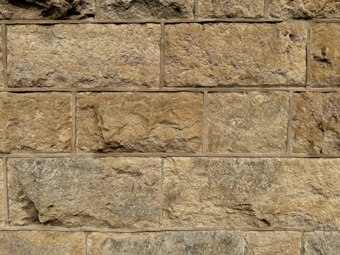A close-up view of a stone wall with large, rectangular blocks. The stones are unevenly textured, showcasing natural variations in color and form. Each block has a slightly different hue, ranging from light tan to deeper brown, with visible mortar lines between the stones.