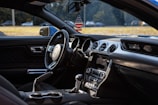 Inside the interior of a car, featuring a steering wheel with a prominent logo, dashboard controls, and air vents. A decorative air freshener in the shape of an American flag hangs from the rearview mirror, and the side view mirror is visible. Sunlight filters through, highlighting the sleek design and materials used in the interior.
