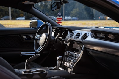Inside the interior of a car, featuring a steering wheel with a prominent logo, dashboard controls, and air vents. A decorative air freshener in the shape of an American flag hangs from the rearview mirror, and the side view mirror is visible. Sunlight filters through, highlighting the sleek design and materials used in the interior.