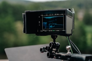 A camera monitor displaying a film scene, mounted on a tripod or stand with a blurred nature background in the distance.