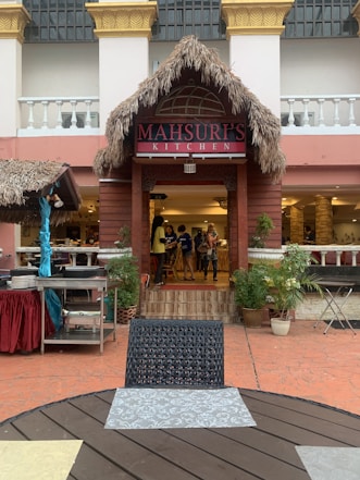 A restaurant entrance with a thatched roof and a sign that reads 'Mahsuri's Kitchen'. In front of the entrance, there is an outdoor dining area with a round table and a woven chair. Several potted plants are placed near the entrance, and a few people are seen standing inside the restaurant. The building has a Mediterranean architectural style with ornate columns and balustrades.