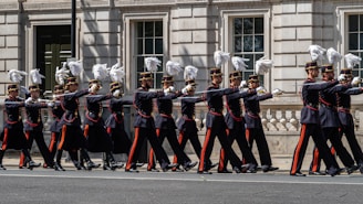 A group of uniformed soldiers marching in a ceremonial procession down a city street. They are wearing dark uniforms with red and gold accents, and their hats are adorned with white plumes. The formation is precise and disciplined, captured against the backdrop of a historic building with large windows and ornate details.