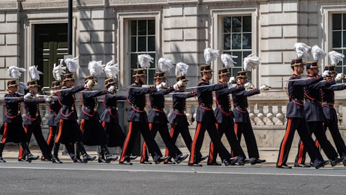 A group of uniformed soldiers marching in a ceremonial procession down a city street. They are wearing dark uniforms with red and gold accents, and their hats are adorned with white plumes. The formation is precise and disciplined, captured against the backdrop of a historic building with large windows and ornate details.