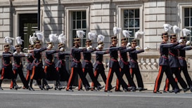 A group of uniformed soldiers marching in a ceremonial procession down a city street. They are wearing dark uniforms with red and gold accents, and their hats are adorned with white plumes. The formation is precise and disciplined, captured against the backdrop of a historic building with large windows and ornate details.