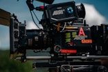A professional film camera with various components and labels is mounted on a rig. The camera includes a lens marked with '85 T1.5', and several sticky notes and labels are visible, including 'ARRE' and 'A005'. The background shows a blurred outdoor scene with blue sky and clouds.