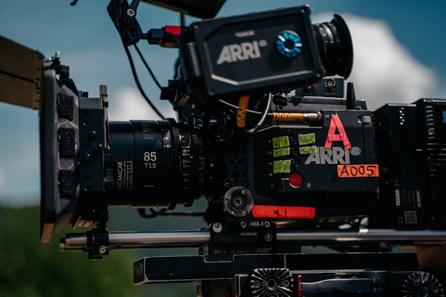 A professional film camera with various components and labels is mounted on a rig. The camera includes a lens marked with '85 T1.5', and several sticky notes and labels are visible, including 'ARRE' and 'A005'. The background shows a blurred outdoor scene with blue sky and clouds.