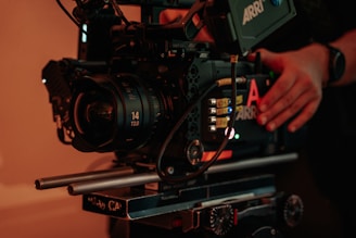 A professional cinema camera setup with a large lens and various controls. The camera is mounted on a rig with visible metal rods and adjustment knobs. There are hands adjusting the camera, suggesting someone is preparing it for filming.