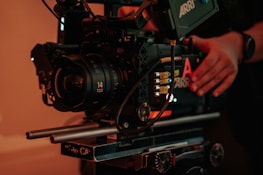 A professional cinema camera setup with a large lens and various controls. The camera is mounted on a rig with visible metal rods and adjustment knobs. There are hands adjusting the camera, suggesting someone is preparing it for filming.