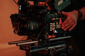 A professional cinema camera setup with a large lens and various controls. The camera is mounted on a rig with visible metal rods and adjustment knobs. There are hands adjusting the camera, suggesting someone is preparing it for filming.
