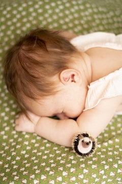 A sleeping baby lies on a patterned green blanket with a small plush hedgehog toy next to them. The baby is wearing a light-colored sleeveless outfit, and their head is turned to the side with their arm tucked under it.