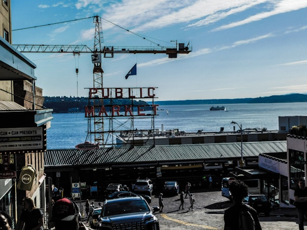 A view of a bustling market area with a prominent sign that reads 'PUBLIC MARKET' in red letters. In the background, there is a large body of water with a few boats and a distant shoreline visible. A construction crane towers over the area. There are various shops and buildings flanking a cobblestone street filled with people and cars.