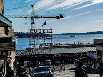 A view of a bustling market area with a prominent sign that reads 'PUBLIC MARKET' in red letters. In the background, there is a large body of water with a few boats and a distant shoreline visible. A construction crane towers over the area. There are various shops and buildings flanking a cobblestone street filled with people and cars.