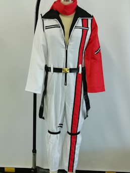 A mannequin is dressed in a white jumpsuit featuring black and red accents. The suit has a prominent vertical red stripe running down the left side, and black stripes on the chest and legs. It is adorned with a gold buckle belt and has a red scarf. A red sleeve contrasts with the otherwise white outfit.
