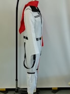A white costume with black accents, including a black belt, is displayed on a mannequin. The costume has a red scarf draped over the shoulders and is set against a plain background. The mannequin is part of a stand with a visible black base and a metal rod that extends vertically.