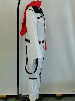 A white costume with black accents, including a black belt, is displayed on a mannequin. The costume has a red scarf draped over the shoulders and is set against a plain background. The mannequin is part of a stand with a visible black base and a metal rod that extends vertically.