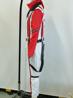 A mannequin displays a full-body suit with a striking color pattern. The suit features red, black, and white colors with bold vertical and diagonal stripes. Its design incorporates a high collar, zippers, and a harness-like strap around the waist and thighs.