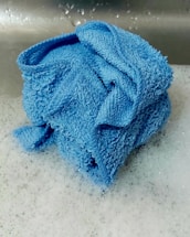 A folded blue towel sits on a layer of soapy bubbles, likely in a sink. The towel's texture is rough, with visible fibers and a saturated blue color. The background shows a metallic surface with water droplets.