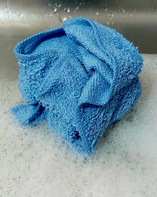 A folded blue towel sits on a layer of soapy bubbles, likely in a sink. The towel's texture is rough, with visible fibers and a saturated blue color. The background shows a metallic surface with water droplets.