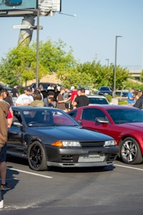 A parking lot filled with people gathering around two cars, one black and one red. A large billboard and trees are visible in the background. The scene seems to capture a casual meet-up or car show with attendees talking and observing the vehicles.
