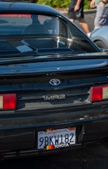The back of a black Toyota MR2 car is parked, featuring a California license plate. In the background, there is a blurred view of two people walking and a silver car.