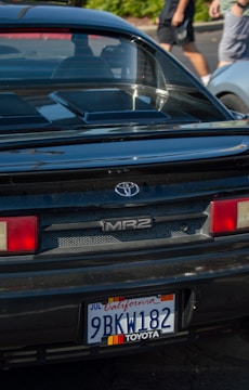 The back of a black Toyota MR2 car is parked, featuring a California license plate. In the background, there is a blurred view of two people walking and a silver car.