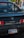 The back of a black Toyota MR2 car is parked, featuring a California license plate. In the background, there is a blurred view of two people walking and a silver car.