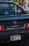 The back of a black Toyota MR2 car is parked, featuring a California license plate. In the background, there is a blurred view of two people walking and a silver car.