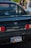 The back of a black Toyota MR2 car is parked, featuring a California license plate. In the background, there is a blurred view of two people walking and a silver car.