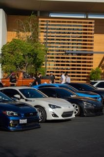 Several sports cars are parked in an orderly array on a paved surface in front of a modern building with wooden accents. A group of people is gathered nearby, possibly attending a car event. Trees provide greenery, complementing the otherwise urban setting.