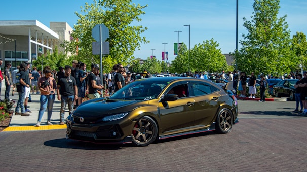 A sleek, metallic brown car with custom modifications attracts a crowd in an outdoor setting. People are gathered around, some taking photos and others simply observing. The background features greenery and buildings, suggesting a car show or event.