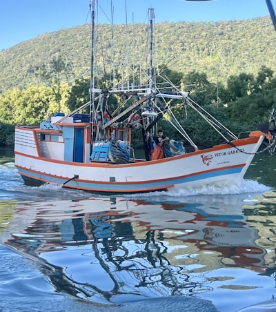 A colorful fishing boat sails along a calm body of water with lush green hills in the background. The boat, named Vitor Gabriel, features vibrant orange and blue accents and intricate rigging. The reflection of the boat creates a visually captivating pattern on the water's surface.
