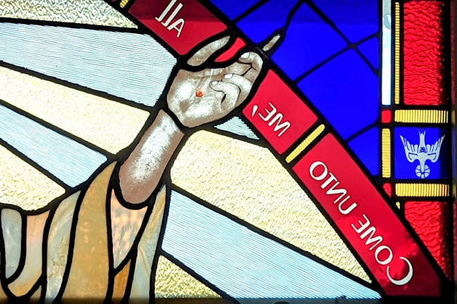 A stained glass window features an outstretched hand with a small red mark on the palm. Surrounding the hand are vibrant colors including blue and red. The words 'COME UNTO ME' are inscribed within a red band. In the corner, there is an image resembling a white dove on a blue background.