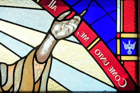 A stained glass window features an outstretched hand with a small red mark on the palm. Surrounding the hand are vibrant colors including blue and red. The words 'COME UNTO ME' are inscribed within a red band. In the corner, there is an image resembling a white dove on a blue background.