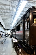 A vintage train on display in a modern, well-lit interior space with high ceilings. The train is dark brown with clean lines and detailed craftsmanship. People are visible in the background, suggesting a museum setting. The sleek design and lighting fixtures enhance the elegance of the space.