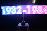 A brightly lit digital display features the years 1982-1984 in neon-style white and blue on the left half and pink on the right half. Below the display, a shiny trophy is placed on a table, silhouetted against the light. The background appears dark to emphasize the glowing text and the reflective surface of the trophy.
