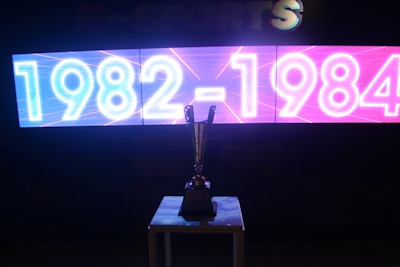 A brightly lit digital display features the years 1982-1984 in neon-style white and blue on the left half and pink on the right half. Below the display, a shiny trophy is placed on a table, silhouetted against the light. The background appears dark to emphasize the glowing text and the reflective surface of the trophy.