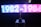 A brightly lit digital display features the years 1982-1984 in neon-style white and blue on the left half and pink on the right half. Below the display, a shiny trophy is placed on a table, silhouetted against the light. The background appears dark to emphasize the glowing text and the reflective surface of the trophy.
