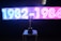 A brightly lit digital display features the years 1982-1984 in neon-style white and blue on the left half and pink on the right half. Below the display, a shiny trophy is placed on a table, silhouetted against the light. The background appears dark to emphasize the glowing text and the reflective surface of the trophy.